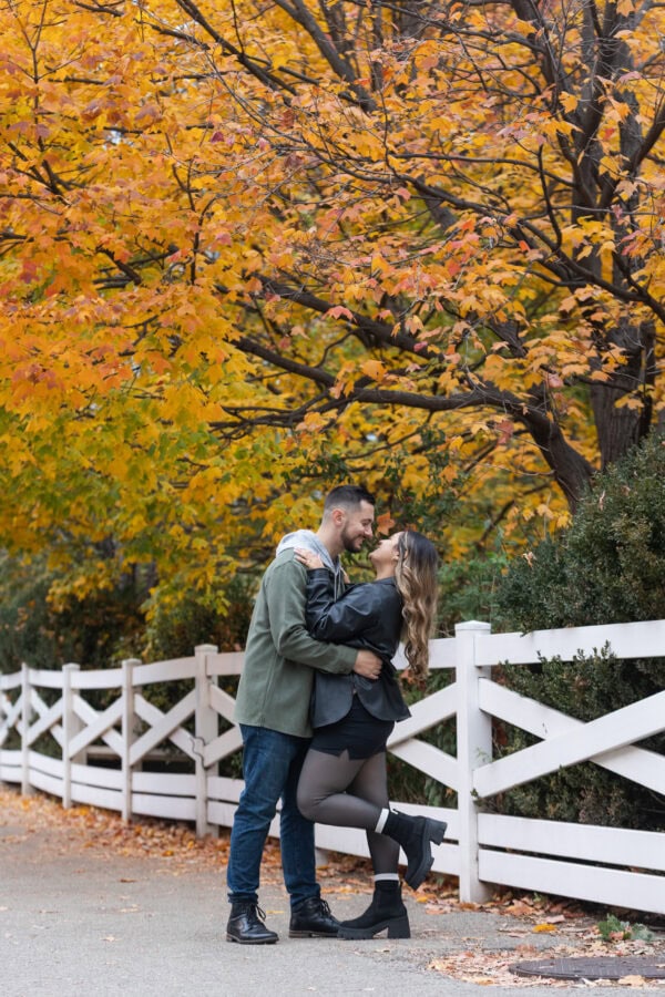 A newly engaged couple hugs in front of colorful fall trees and white fence in Chicagos Lincoln Park