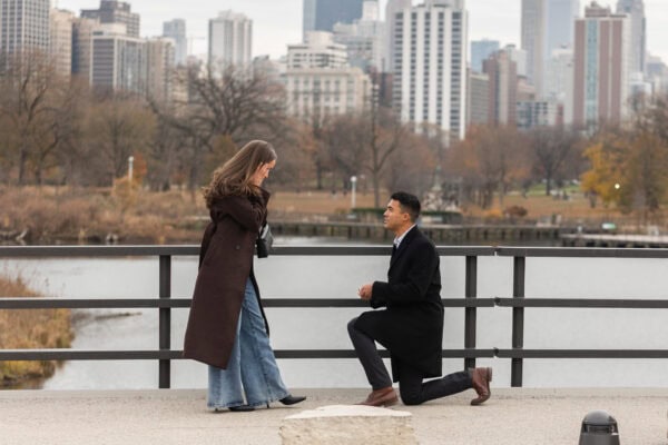 Man kneels to propose by the water as she holds her face, with Chicago skyline behind them during a surprise proposal