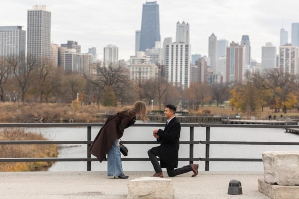 Man kneels to propose by the water as partner leans in, with Chicago skyline behind them during a surprise proposal