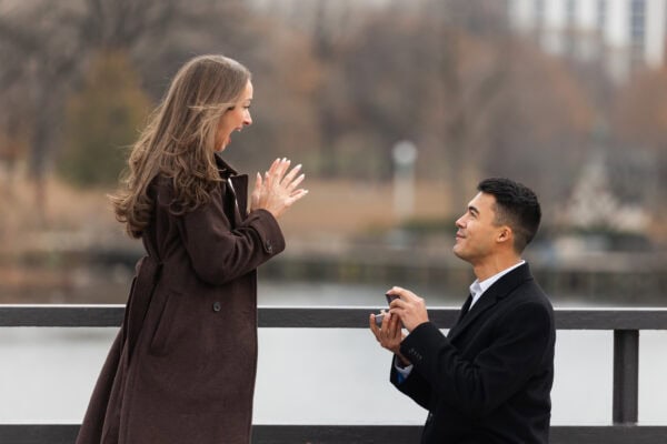 Man kneels to propose by the water as partner screams with joy with Chicago skyline behind them during a surprise proposal