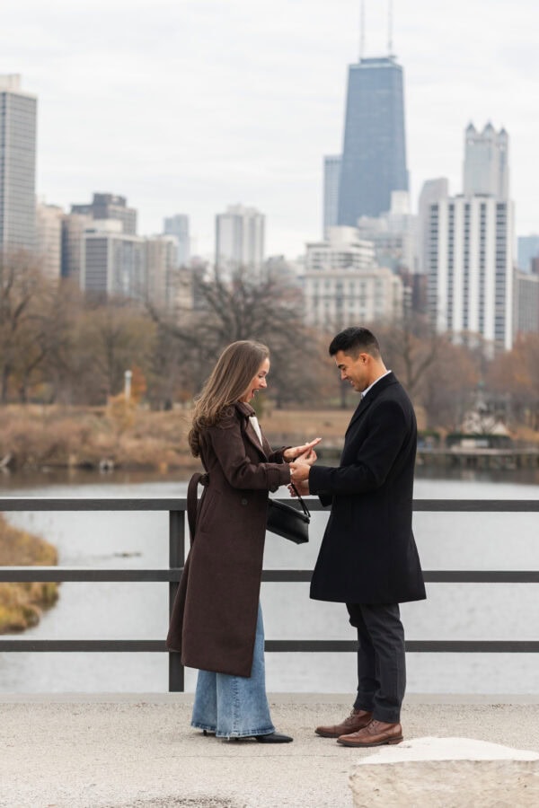 Couple looks at the new engagement ring on her finger Chicago skyline behind them during a surprise proposal