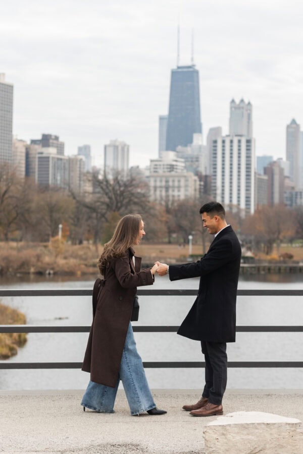 Couple holds hands and celebrates with Chicago skyline behind them during a surprise proposal