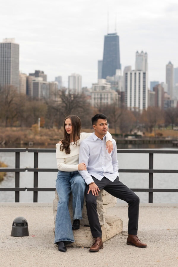 Couple poses for a photo during an engagement session seated on a rock in front of the Chicago skyline