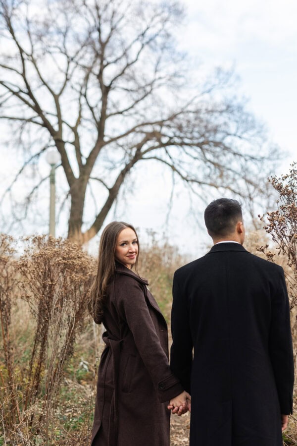 Couple poses for a photo during an engagement session as she looks back at the camera surrounded by trees