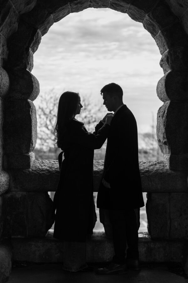 black and white silhouette of a woman adjusting a mans tie under a stone arch entryway