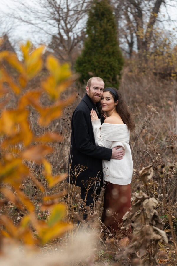 a couple poses for an engagement photo as they look straight at the camera while standing in an autumn colored field