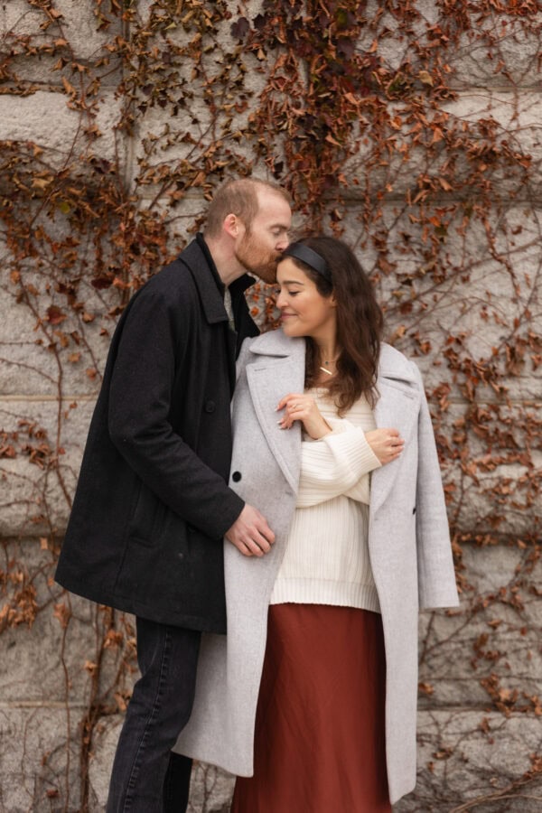 a couple poses for an engagement photo as the man kisses his fiance on the forehead in front a stone wall of red ivy