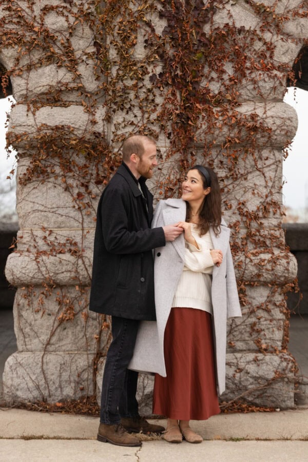a couple poses for an engagement photo as they hold hands in front a stone wall of red ivy