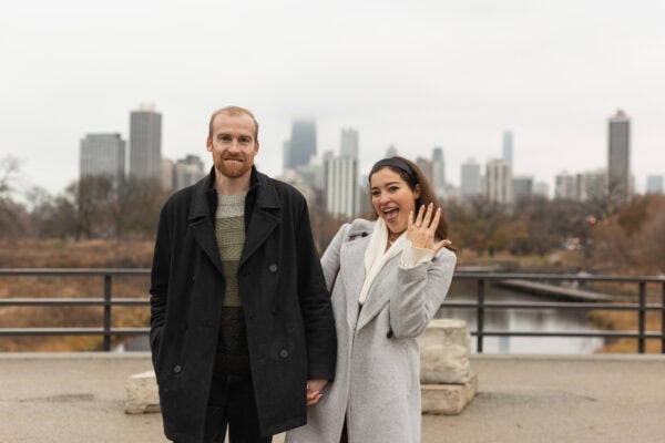 a newly engaged couple looks at the camera and smiles, while the fiance shows off her new ring