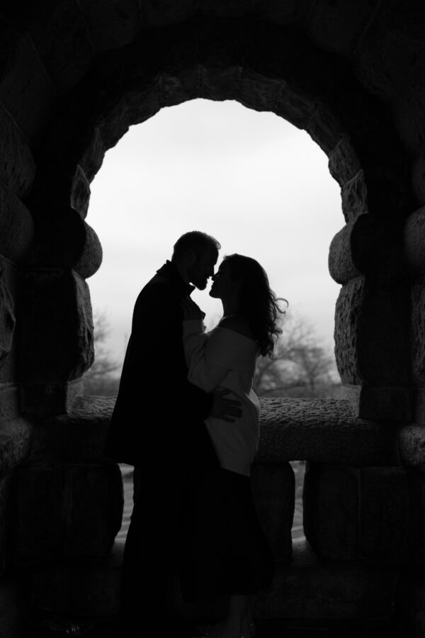 an engaged couple stands nose to nose looking into each others eyes standing under a stone arch
