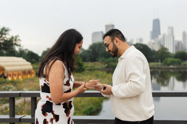 a man places an engagement ring on his fiances hand after proposing to her on a bridge in front of the chicago skyline at lincoln park zoo