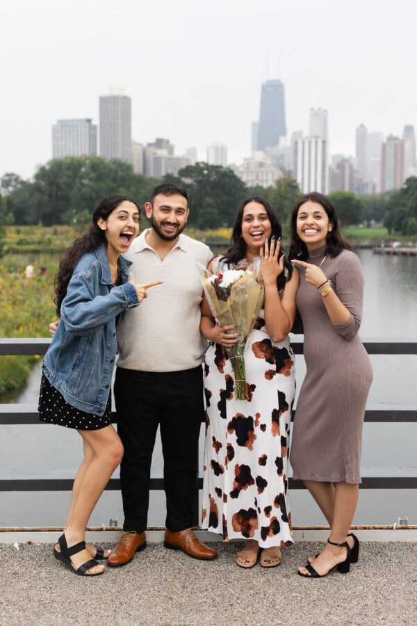 a group of friends poses for a photo in front of the chicago skyline while pointing to the engagement ring on their friends hand