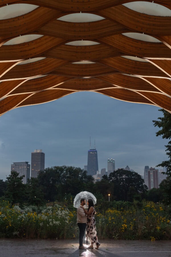 a couple poses for a photo at night under an umbrella in front of the chicago skyline
