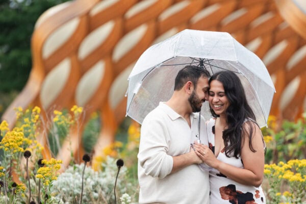 a newly engaged couple poses for a photo under an umbrella in the rain at lincoln park zoo standing in a field of flowers