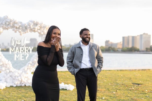 a newly engaged couple has faces of surprise in Chicago in front of a hearts shaped installation made of white roses with a sign that reads "will you marry me"