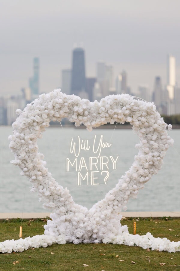 a installation of a heart shaped rose structure with a neon light display reading "will you marry me" is displayed in front of the Chicago Skyline on Montrose harbor