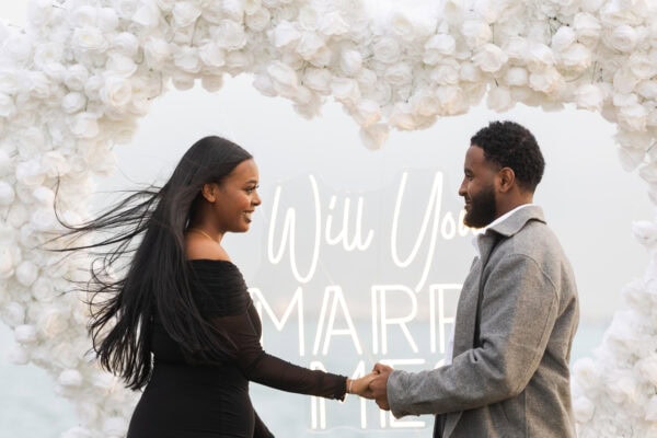 a man proposes to his girlfriend in Chicago in front of a hearts shaped installation made of white roses with a sign that reads "will you marry me"