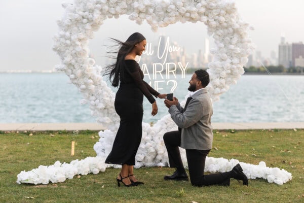 a man is down on one knee proposing to his girlfriend in Chicago in front of a hearts shaped installation made of white roses with a sign that reads "will you marry me"