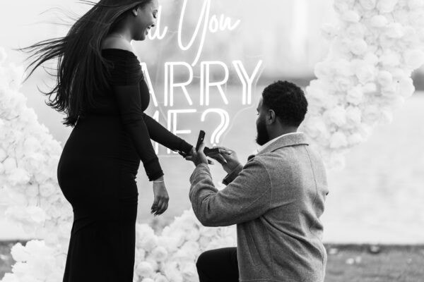a man proposes to his girlfriend by placing a diamond ring on her finger in Chicago in front of a hearts shaped installation made of white roses with a sign that reads "will you marry me"