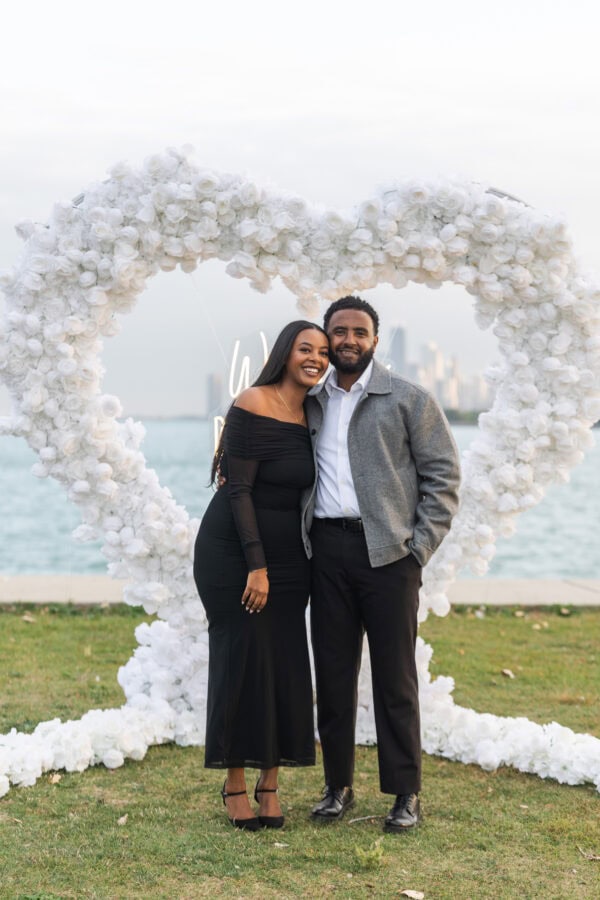 a newly engaged couple poses for a photo in Chicago in front of a hearts shaped installation made of white roses with a sign that reads "will you marry me"