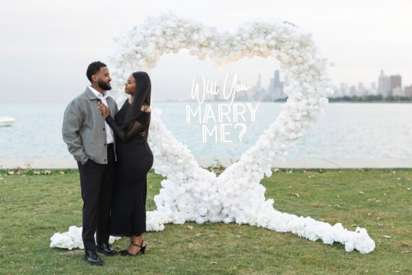 a newly engaged couple poses for a photo in Chicago in front of a hearts shaped installation made of white roses with a sign that reads "will you marry me"