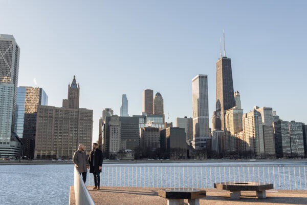 Couple holding hands by the lake with Chicago skyline behind them, posing for an engagement photo