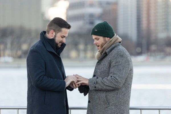 Close-up of one man placing an engagement ring on his partner’s finger by the lake during a Chicago surprise proposal
