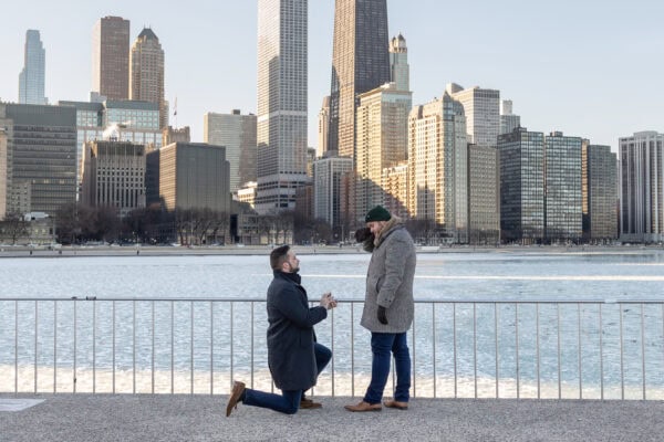 Man kneels to propose by the lake with Chicago skyline behind them as partner reacts with surprise