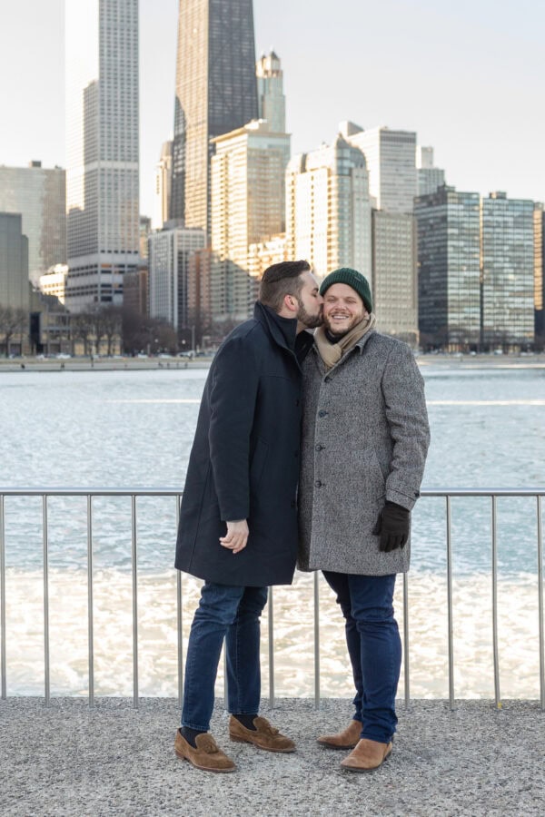 One man kisses his partner’s cheek by the lake with Chicago skyline behind them, celebrating their surprise proposal