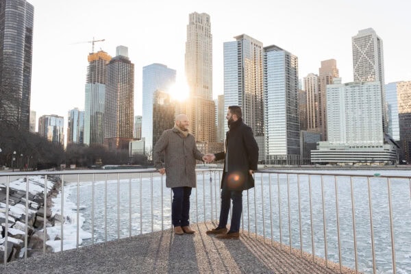 Couple sitting together by the lake at sunset with Chicago skyline behind them, posing for an engagement photo