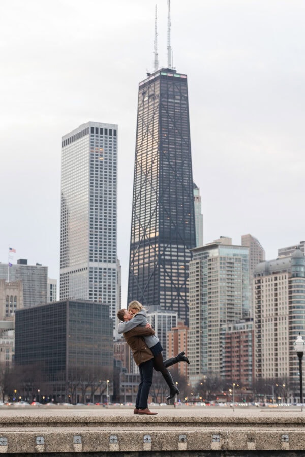 Newly engaged couple celebrates with a joyful embrace at Olive Park with Chicago's skyline behind them.