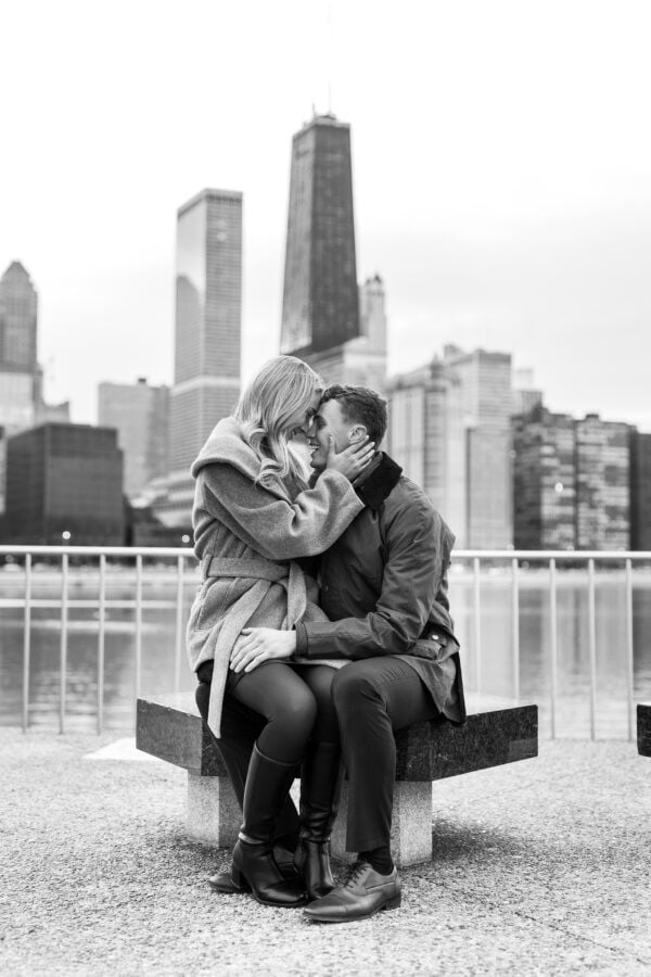 Engaged couple cuddles on a bench beside Lake Michigan with Chicago's skyline behind them.