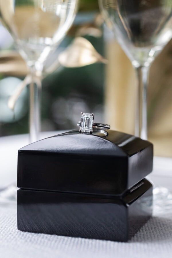 a diamond engagement ring sits on top of its black box in front of two glasses of champagne
