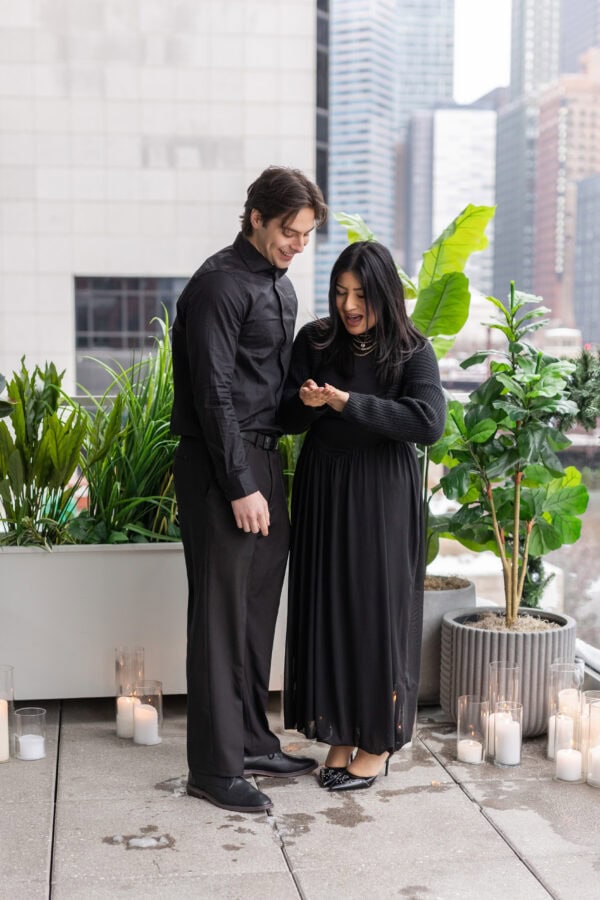 a man and his girlfriend look at her engagement ring on her hand just moments after he proposed to her as they stand on the RPM terrace on the chicago river