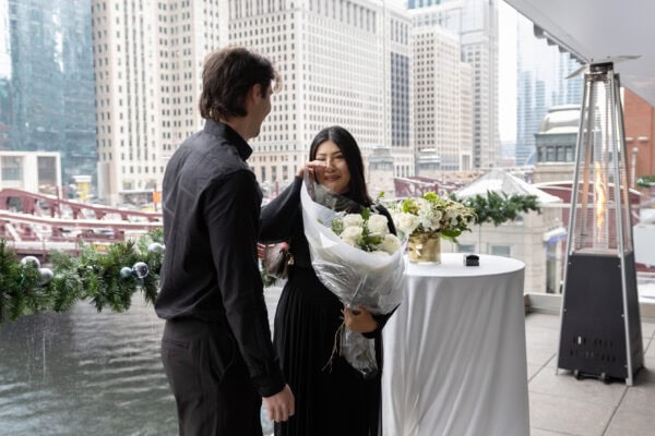 a man looks at his girlfriend as she wipes a tear of happiness off her cheek as she holds a bouquet of white roses just moments after she said yes to her boyfriends proposal
