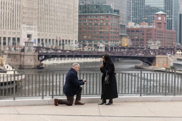 a man gets down on his knee as he proposes to his girlfriend by using a card trick while they stand in front of the chicago downtown skyline at River Point Park