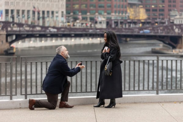 a man gets down on his knee as he proposes to his girlfriend by using a card trick while they stand in front of the chicago downtown skyline at River Point Park