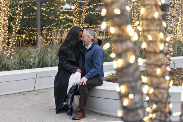 a couple poses for a photo just after getting engaged. They sit surrounded by trees covered in white christmas lights