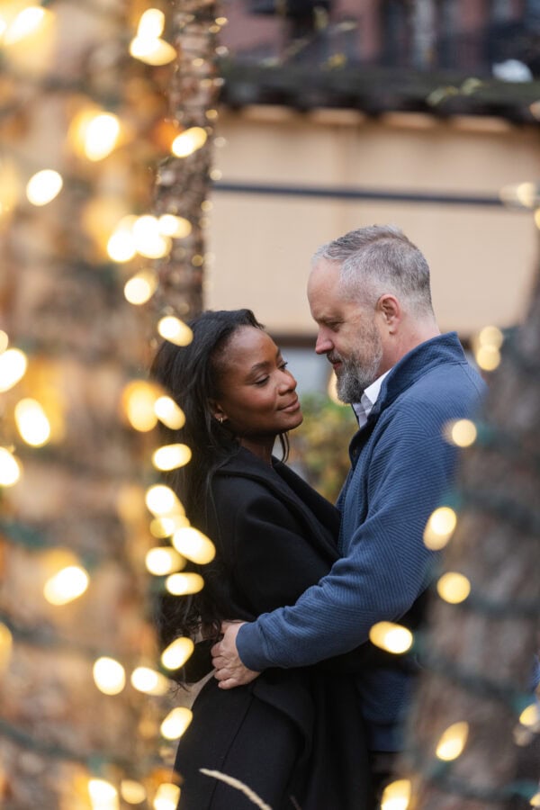 a couple poses for a photo after just getting engaged. They are surrounded my trees covered with christmas lights