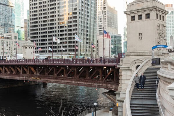 Couple walking down Chicago Riverwalk steps with skyline behind them, posing for an engagement photo