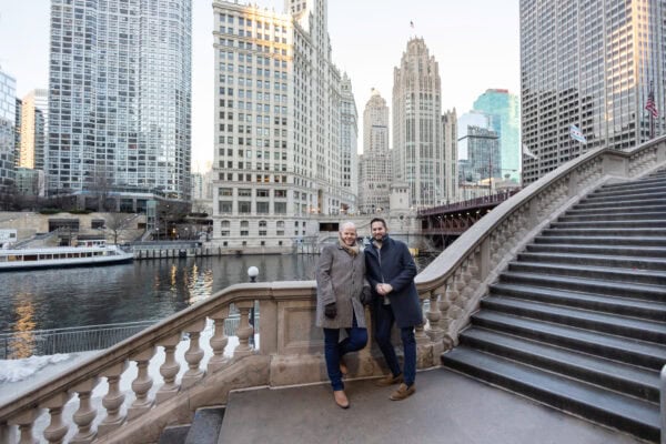 Couple smiling on Chicago Riverwalk steps with skyline behind them, posing for an engagement photo