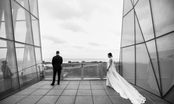 Bride and groom stand on a terrace with glass walls, overlooking a landscape. Bride in a long dress; groom in a suit with his back turned. Black and white image.
