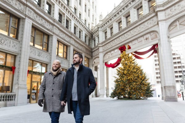A same sex couple poses for an engagement photo at the Wrigley Building in Chicago with Lit Christmas Tree